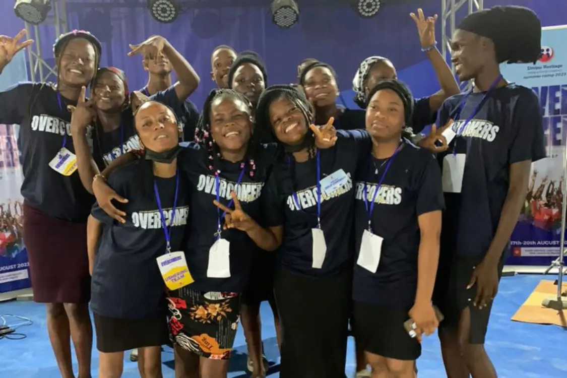 A group of smiling young women wearing matching “OVERCOMERS” T-shirts and name tags pose together indoors, some making peace signs and cheerful gestures, standing on a blue floor with a vibrant backdrop behind them.