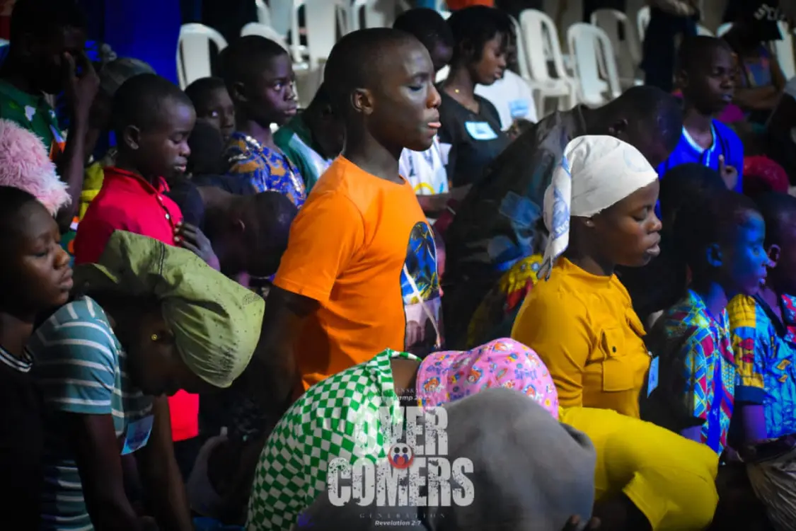 A group of children, some standing and some kneeling, are gathered indoors with heads bowed in prayer or reflection. The scene is colorful, with vibrant clothing and white chairs in the background. The words OVERCOMERS are visible at the bottom.