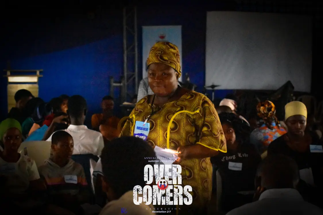 A woman in patterned yellow attire stands among seated people at an indoor event. She holds papers and wears a badge. The background features a dark blue wall, and OVERCOMERS is boldly displayed at the bottom.